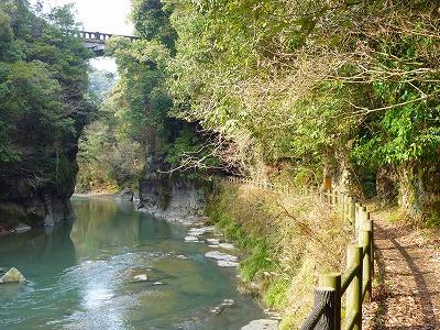 青緑色の川面と陽射しが差す小道、高い位置に橋がかかっている橋満橋周辺の風景写真