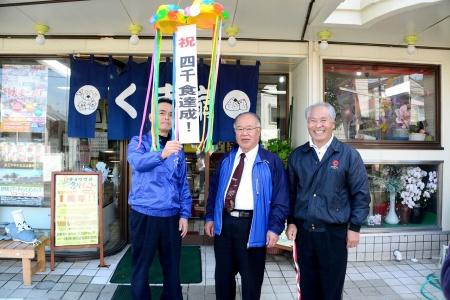ジャンパーを着た男性三名が並んで記念のくす玉を割っている写真