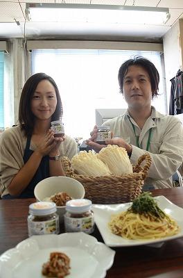 瓶詰の「えのきのしょうゆ炊き」を手にもってパスタやご飯などが並んだテーブルの近くで笑顔を見せる田地さんと瀬尾さんの写真
