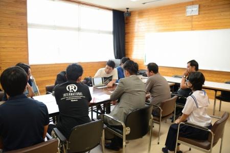 会議室で、小さいパソコンを見せて話している男性と、その画面を周りに座った関係者たちが見ている事前打ち合わせの写真