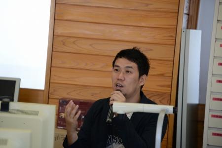 マイクを持った男性が、手を動かしながら座って話している写真