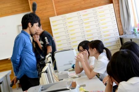 パソコンの前に座って、手を動かしながら話している女子生徒たちと、その様子を立って見ている男性の写真