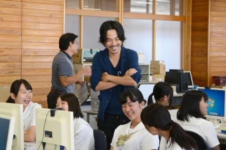 パソコンの前に座っている女子生徒たちと、腕組みをした男性が笑顔を浮かべている写真