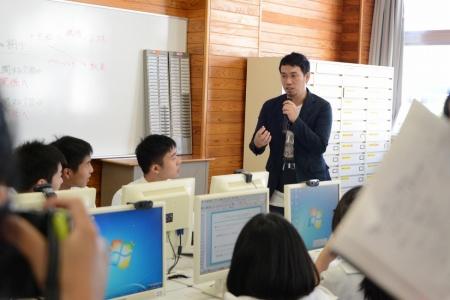 立ってマイクを持って話している男性と、パソコンの前に座って聞いている生徒たちの写真