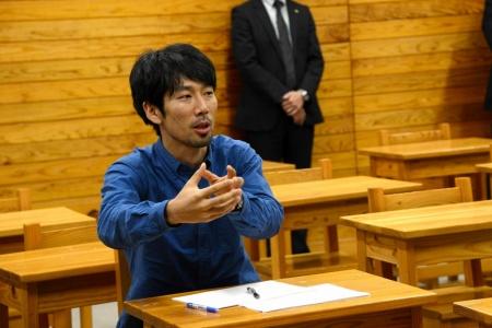 青いシャツを着た男性が、手を動かして説明している写真