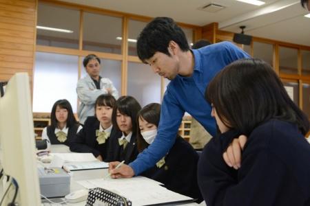 かがみこんで机の上の紙に書き込んでいる男性と、周りに座って聞いている生徒たちの写真