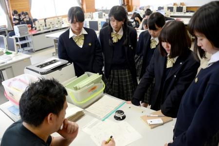 座って絵コンテの紙に書き込んでいる男性と、周りを囲むように立っている女子生徒たちの写真