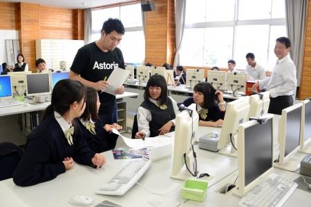 パソコンが並んだ教室で、黒いシャツを着た男性と生徒たちが話し合っている写真