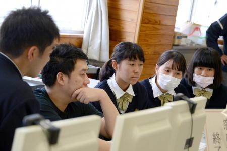 パソコン画面を見つめる黒いシャツを着た男性と、周りに座って同じ画面を見ている生徒たちの写真