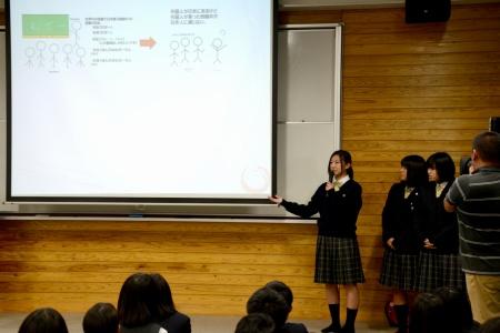 イラストのある資料が表示されたスクリーンの横に女子生徒が立って、説明している写真