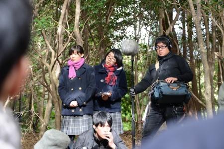 撮影機材を持って立っている男性と、その横に立って撮影を見学している二人の女子生徒たちの写真