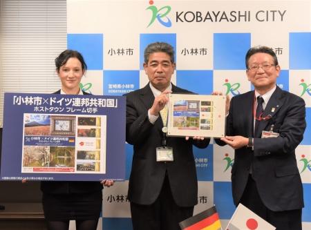 市長とスーツ姿の男女がドイツホストタウンフレーム切手のPRイメージのボードを手に笑顔で立っている写真