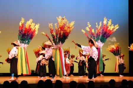 カラフルな造花や装飾を背負い、ステージの上で列を成して舞を披露する演者たちの写真