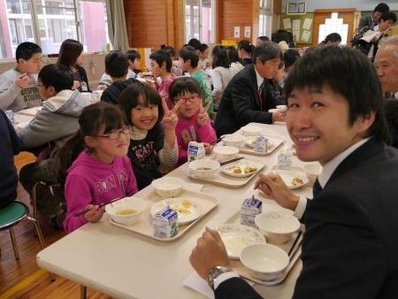 笑顔の子供たちとスーツ姿の大人が一緒になって給食を食べている写真