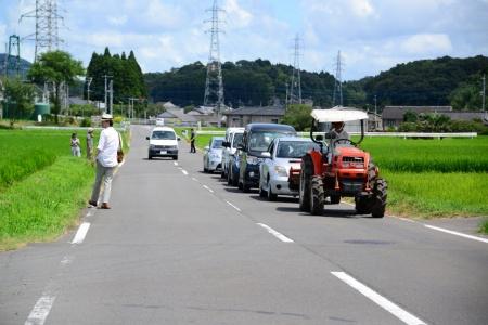 田んぼの隣の車道で、トラクターを先頭に渋滞が起きている写真