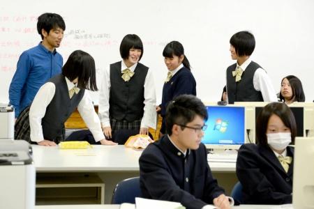 パソコンが並んでいる教室で、生徒たちと男性が集まって笑顔で話し合っている写真