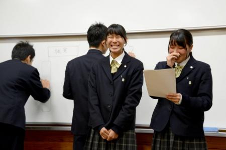 笑顔で並んで立っている女子生徒たちと、後ろでホワイトボードに書き込んでいる男子生徒たちの写真