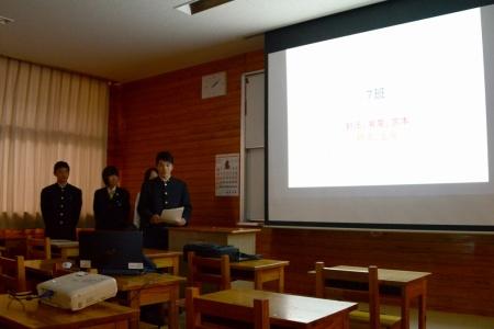 資料が表示されたスクリーンの横に立って、生徒たちがプレゼンテーションをしている写真