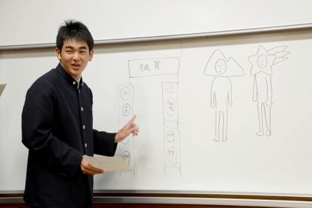 キャラクターが描かれたホワイトボードを指差しして説明している男子生徒の写真