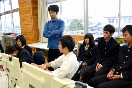 パソコンの周りに座っている生徒たちと、後ろで腕組みをして立っている男性の写真