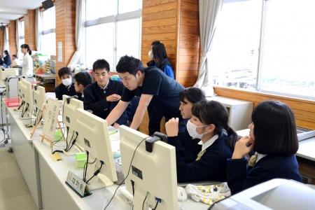 かがみこんでパソコンを操作する黒いシャツを着た男性と、周りに座って見ている生徒たちの写真